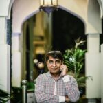 Home A South Asian man in casual attire talks on his mobile phone under a lit archway.